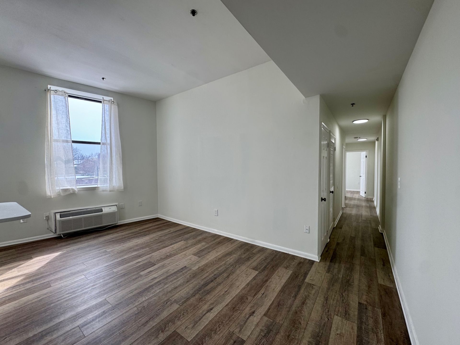 Empty white apartment hallway with wood floors and a windowed room on the left