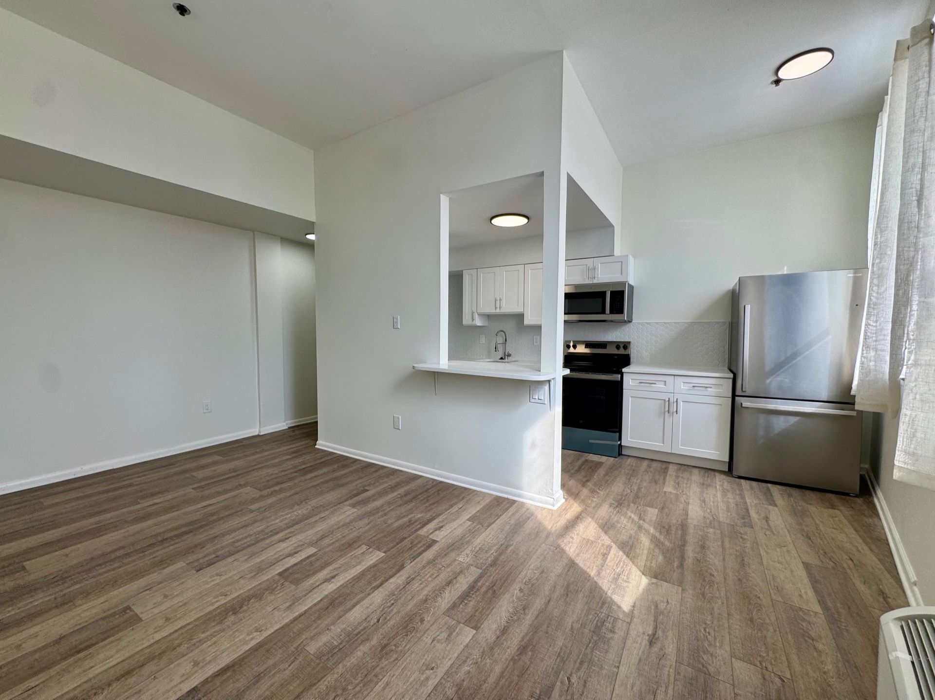 Empty modern apartment kitchen and living area with wood floors, white walls, and stainless steel fridge.