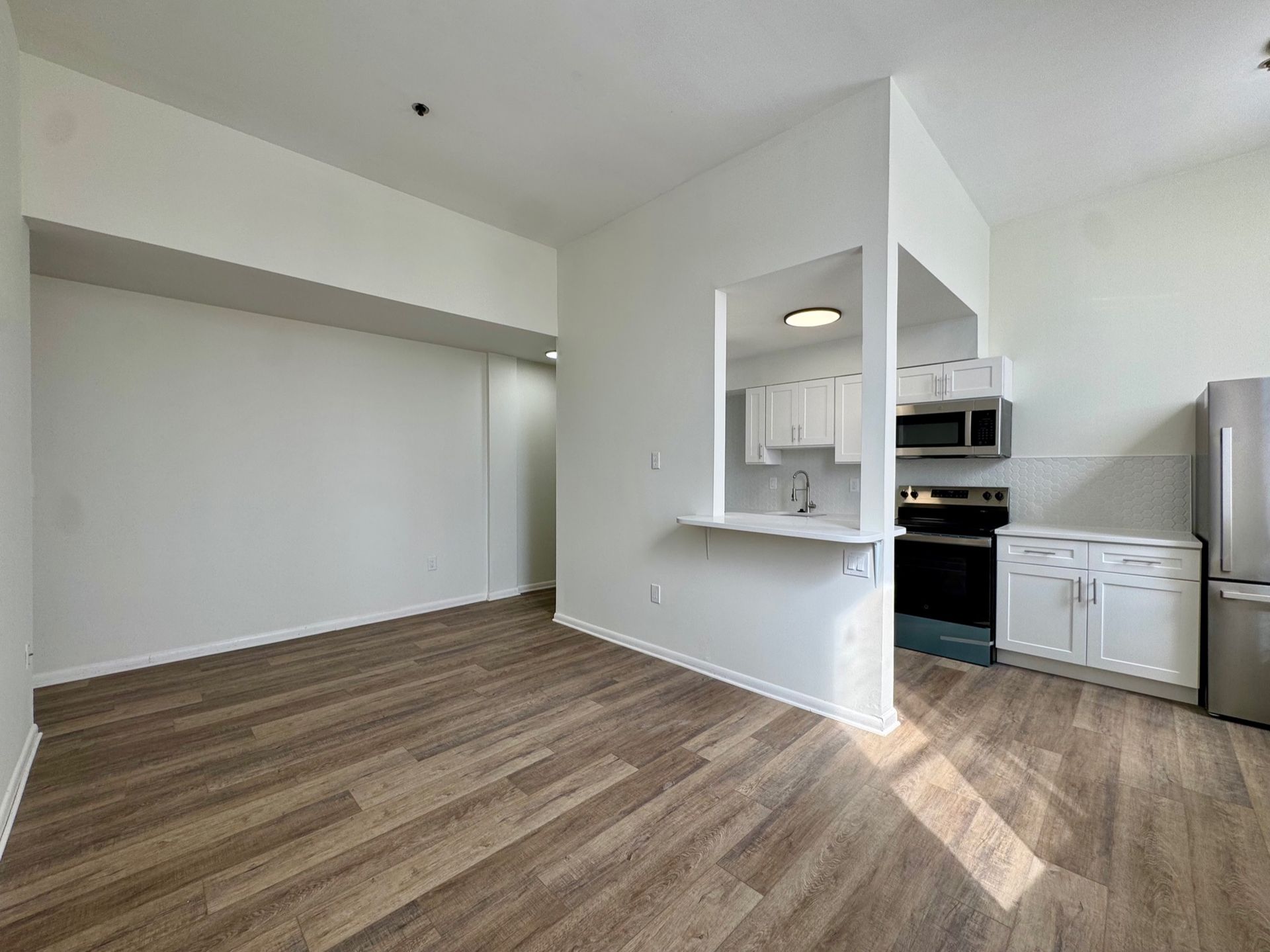Empty modern apartment living area with wood-look floors and open kitchen pass-through.