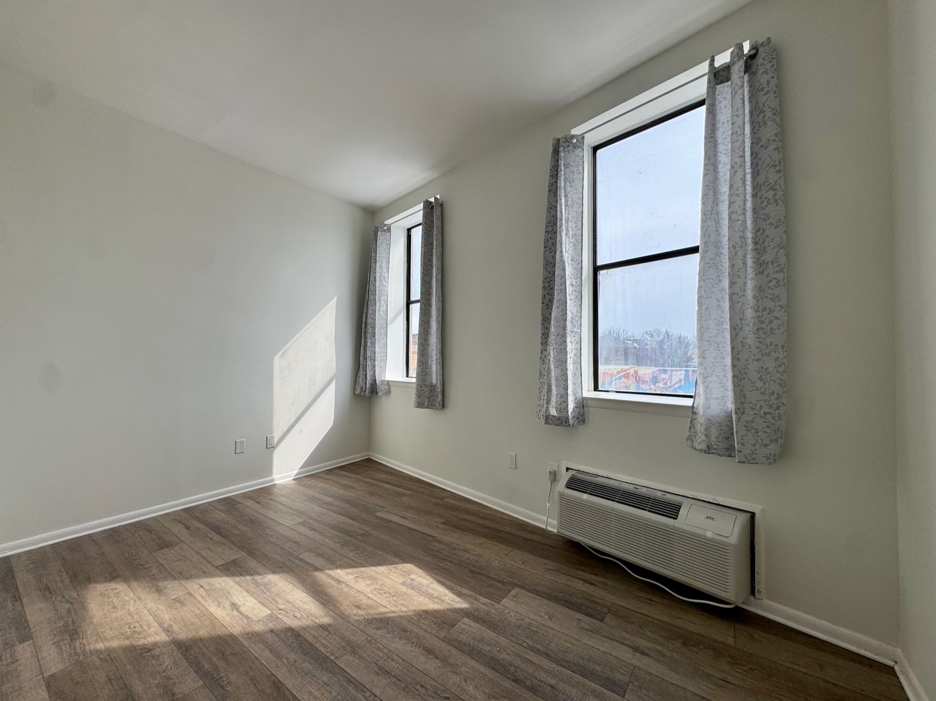 Empty room with wood floor, white walls, two windows, and a wall heater