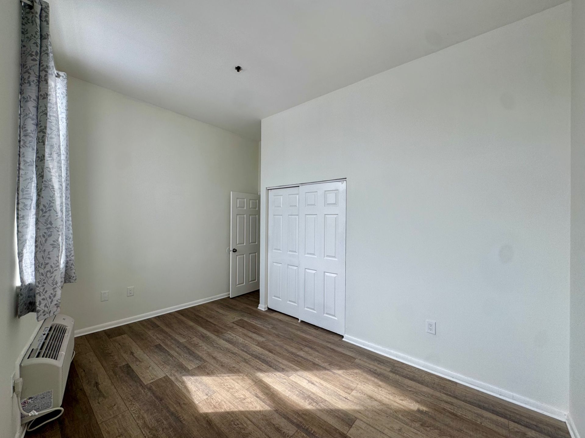 Empty white bedroom with wood floor, window, closet doors, and a wall-mounted heater