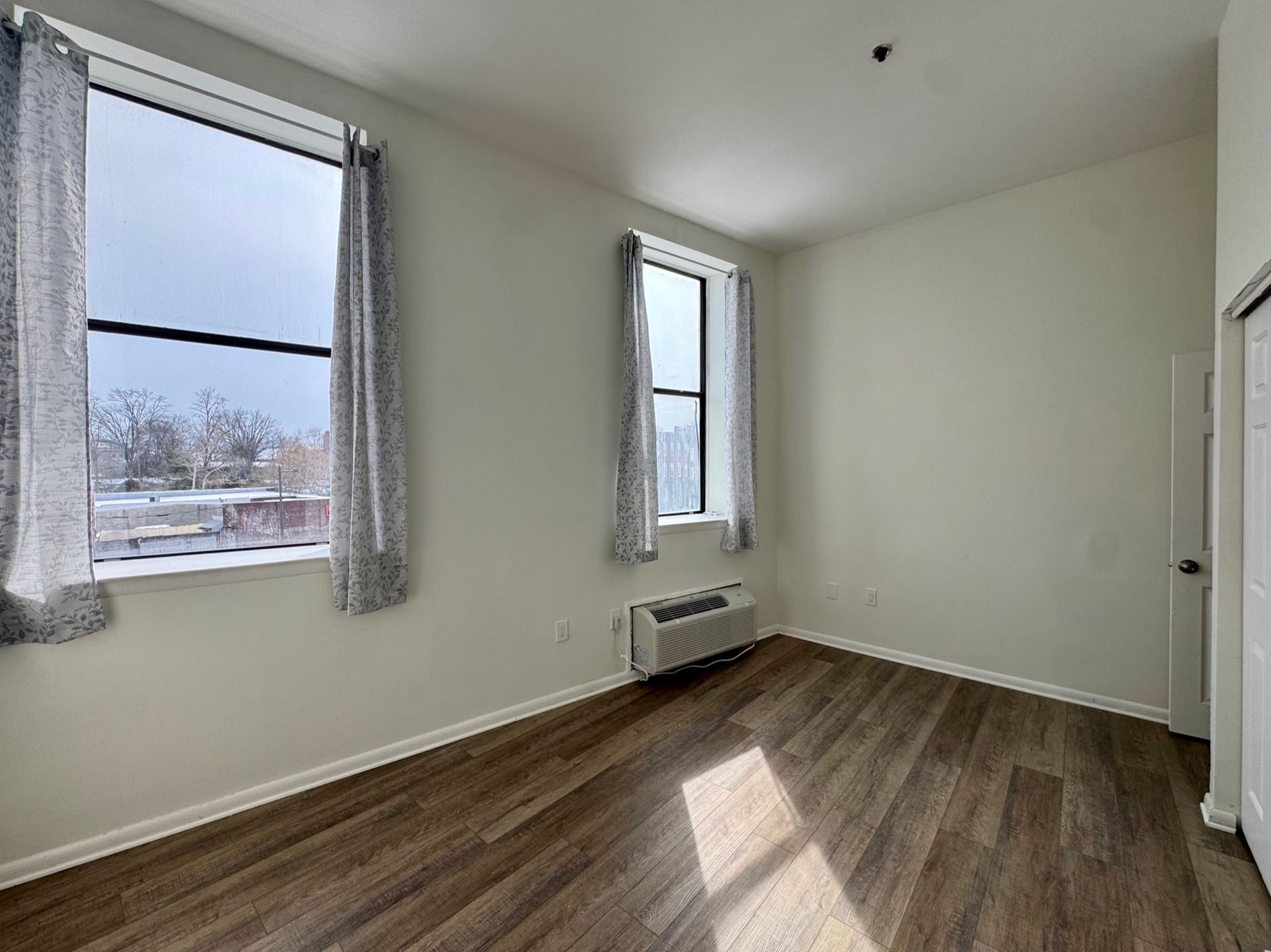 Empty room with wood floor, two windows, and a wall-mounted heater in natural light