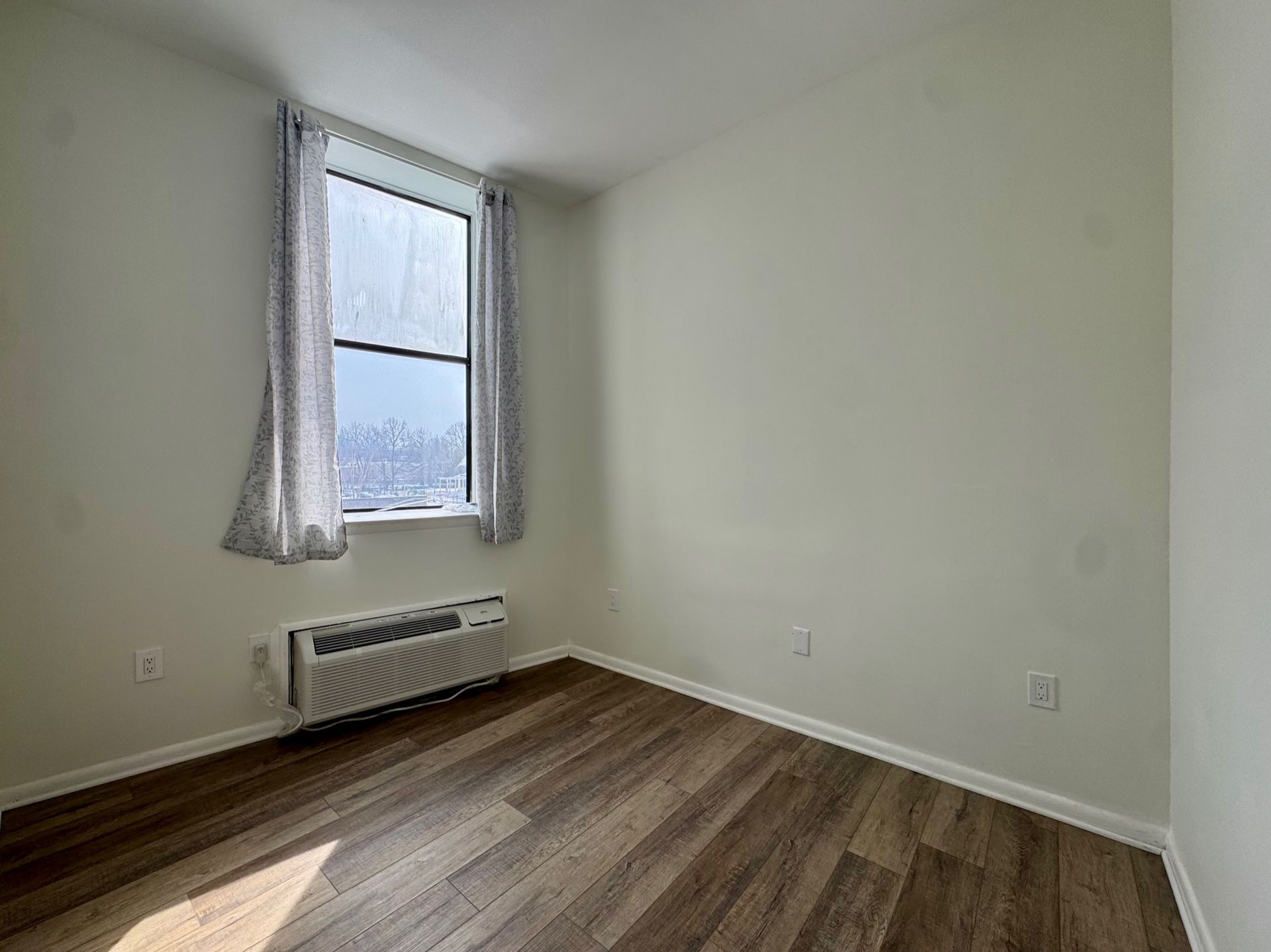 Empty room with wood floors, white walls, a window, and a wall-mounted heater