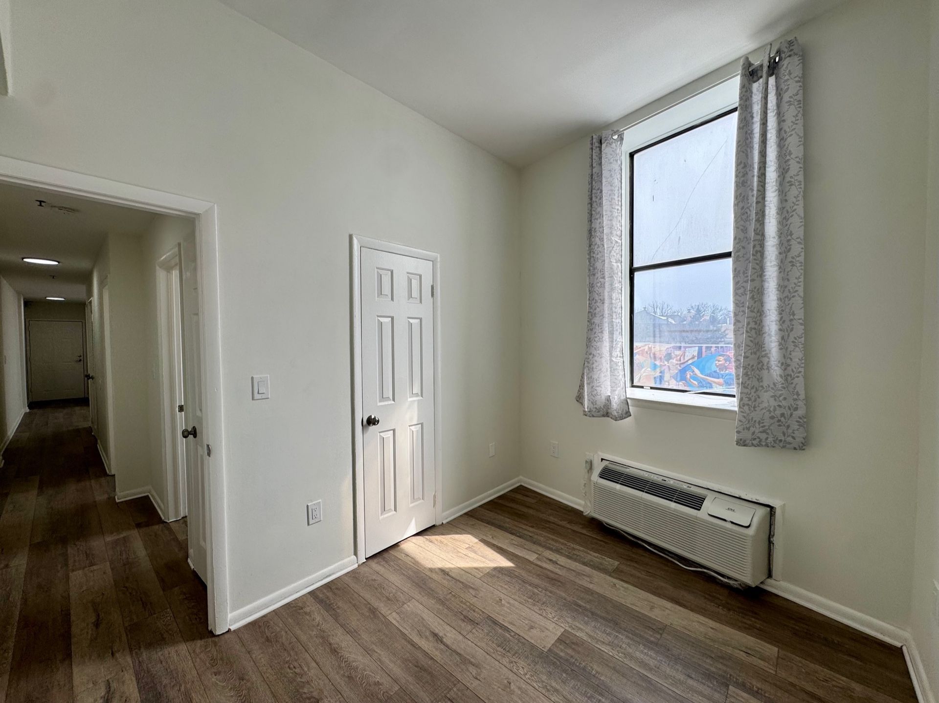 Empty white room with wood floor, tall window, closet door, and hallway opening
