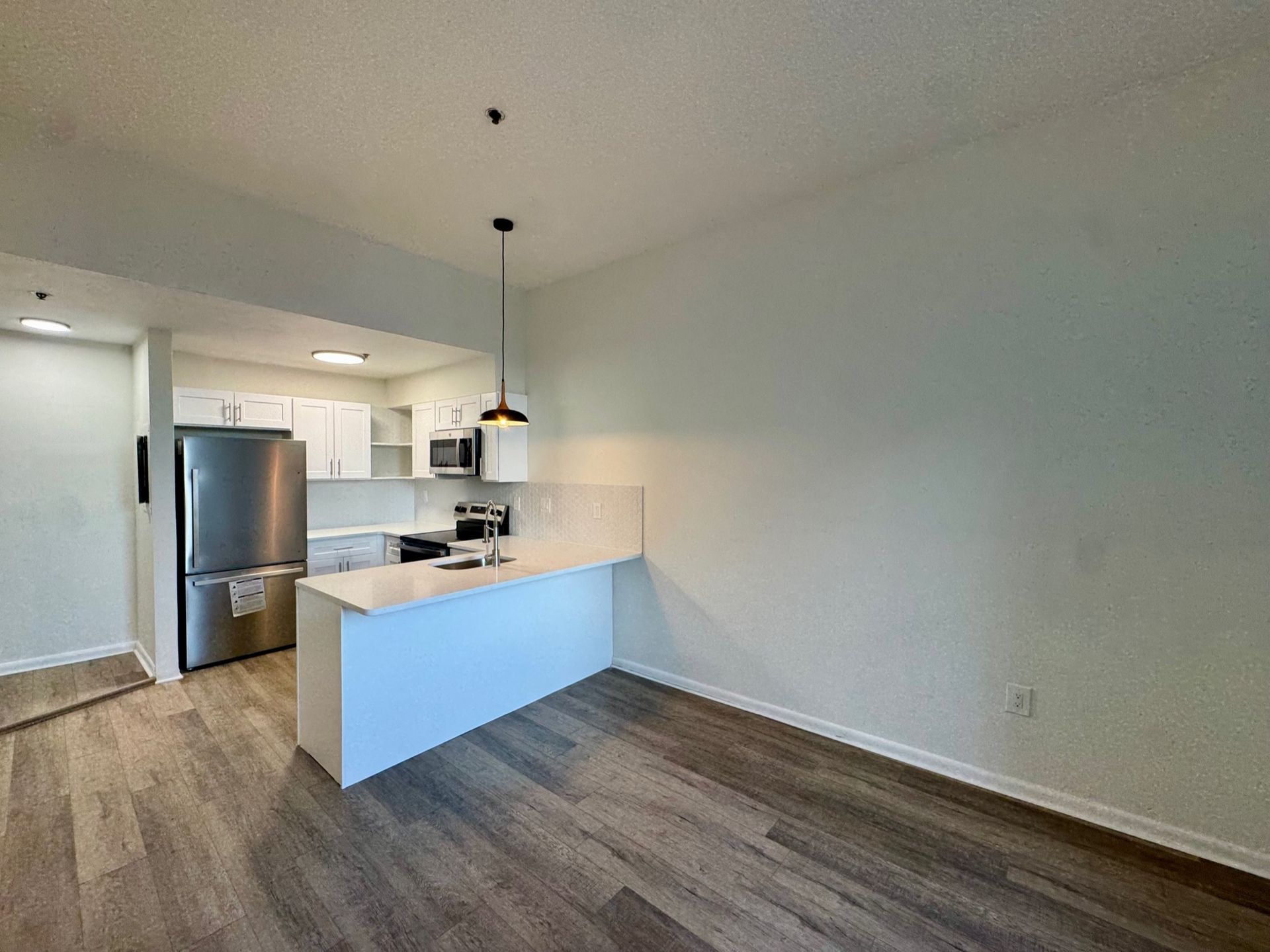 Empty modern kitchen and dining area with stainless steel refrigerator, white island, and wood-look floor.