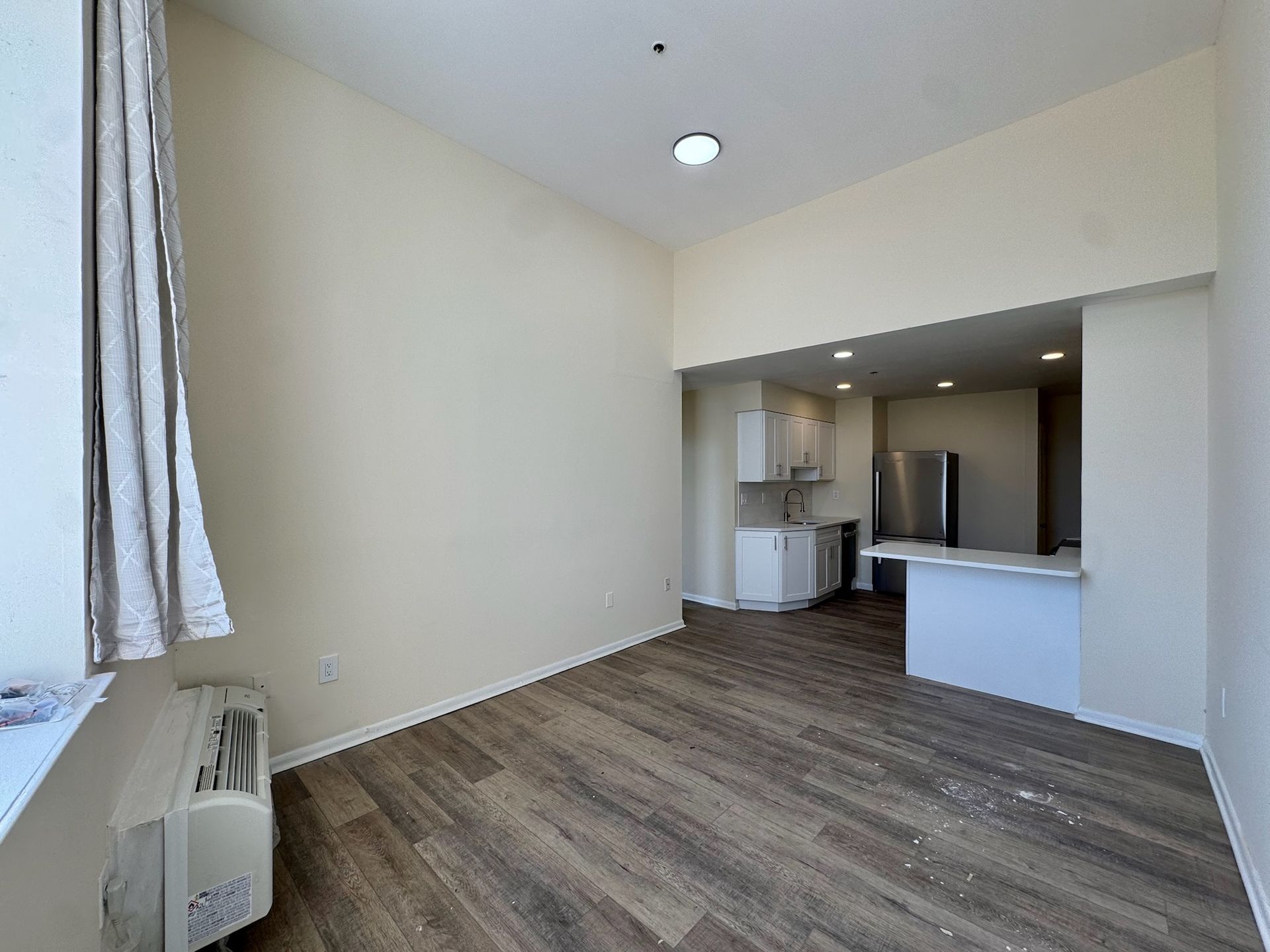 Empty beige living room with wood floors and open kitchen pass-through, lit by a ceiling fixture.