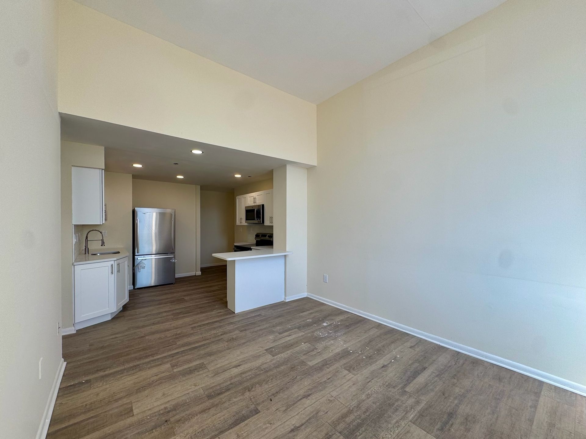 Empty open-plan living room with wood flooring and a small kitchen area with stainless steel fridge.