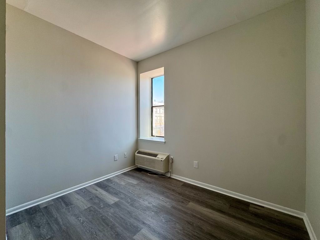 Empty room with gray walls, dark wood floor, and a small window with a wall heater below it