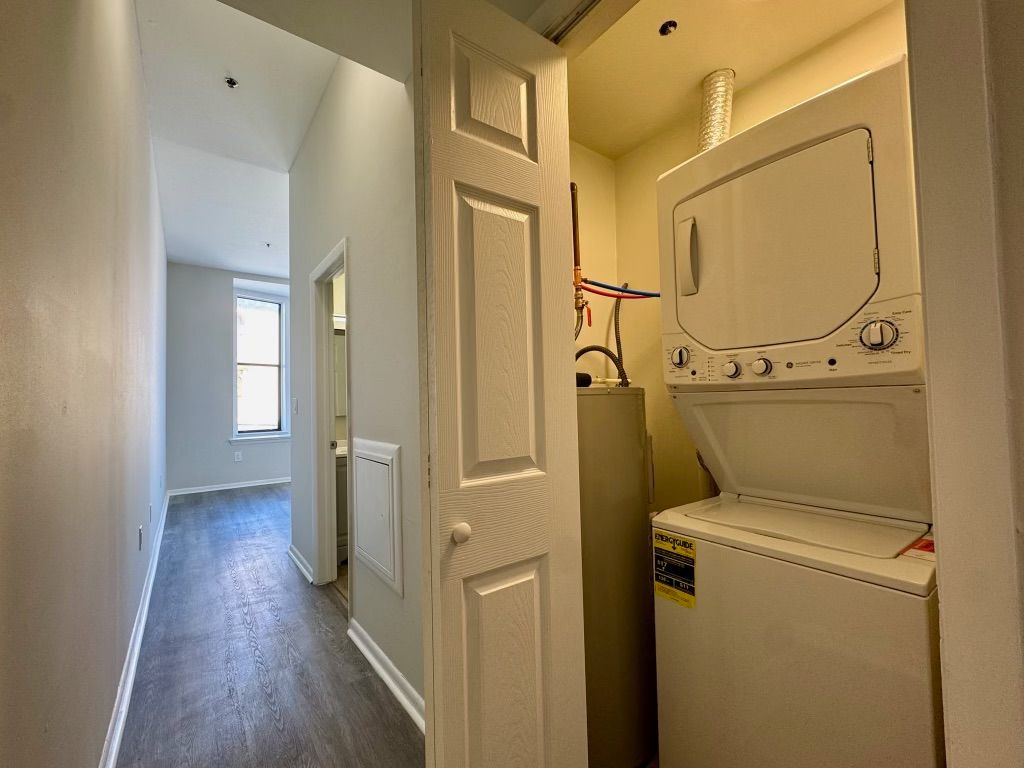 Narrow hallway with laundry closet and stacked washer-dryer units, white walls and wood flooring.