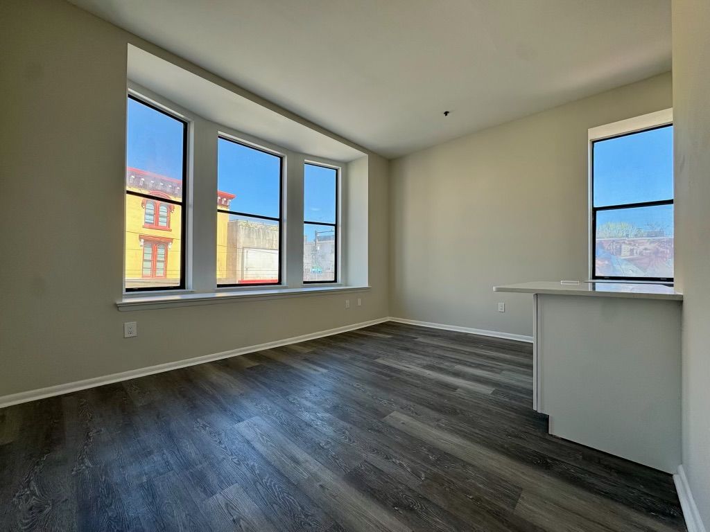Empty modern apartment room with dark wood floors, large windows, and white walls