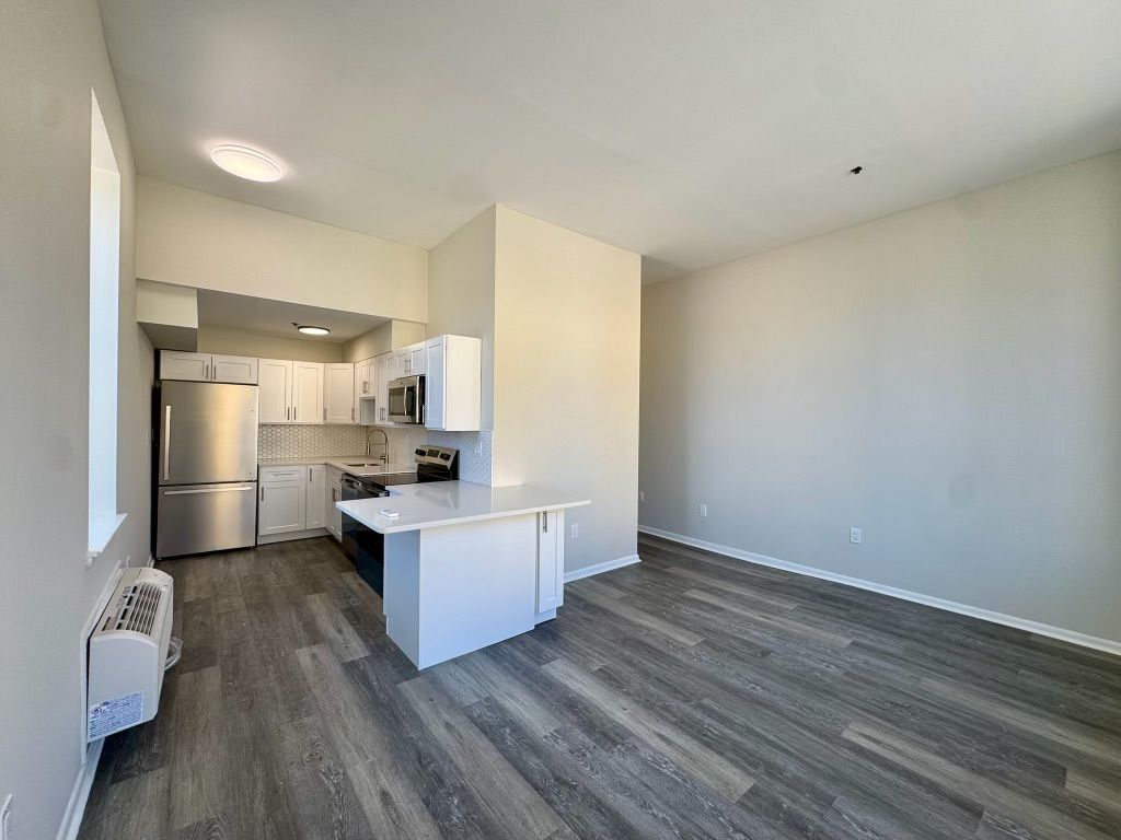 Empty apartment living area with gray floors, white walls, and a compact kitchen with stainless fridge.
