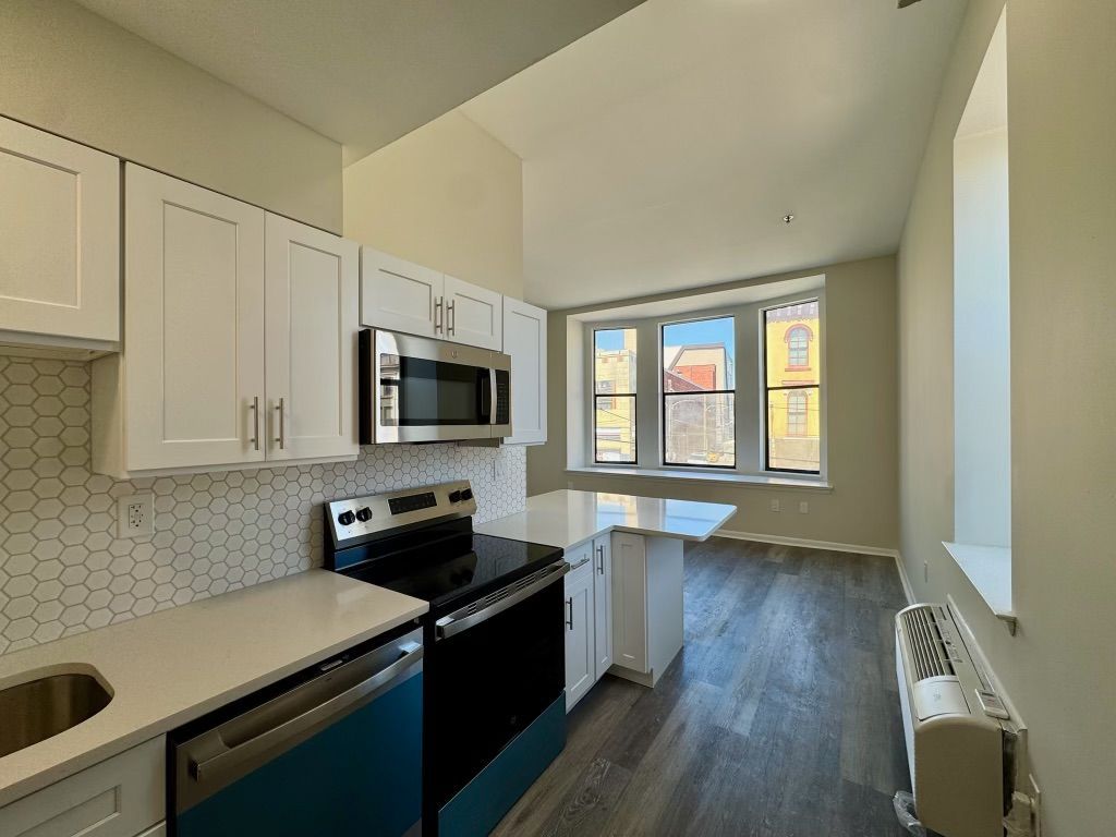 Bright galley kitchen with white cabinets, black stove, and large windows opening to a room with wood floors.
