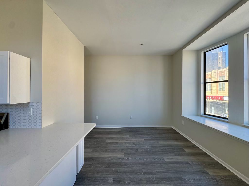 Empty modern kitchen and living area with gray wood floors, white cabinets, and large window.