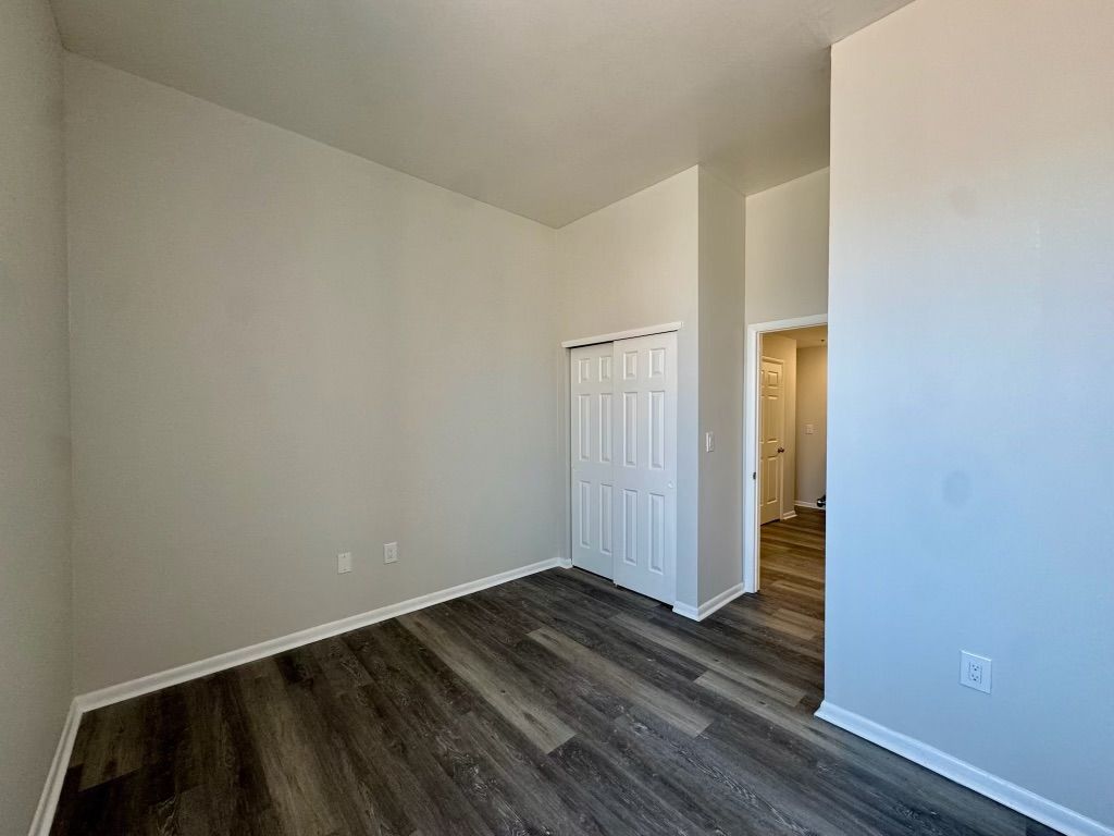 Empty bedroom with dark wood floors, light walls, and open closet doors leading to a hallway.