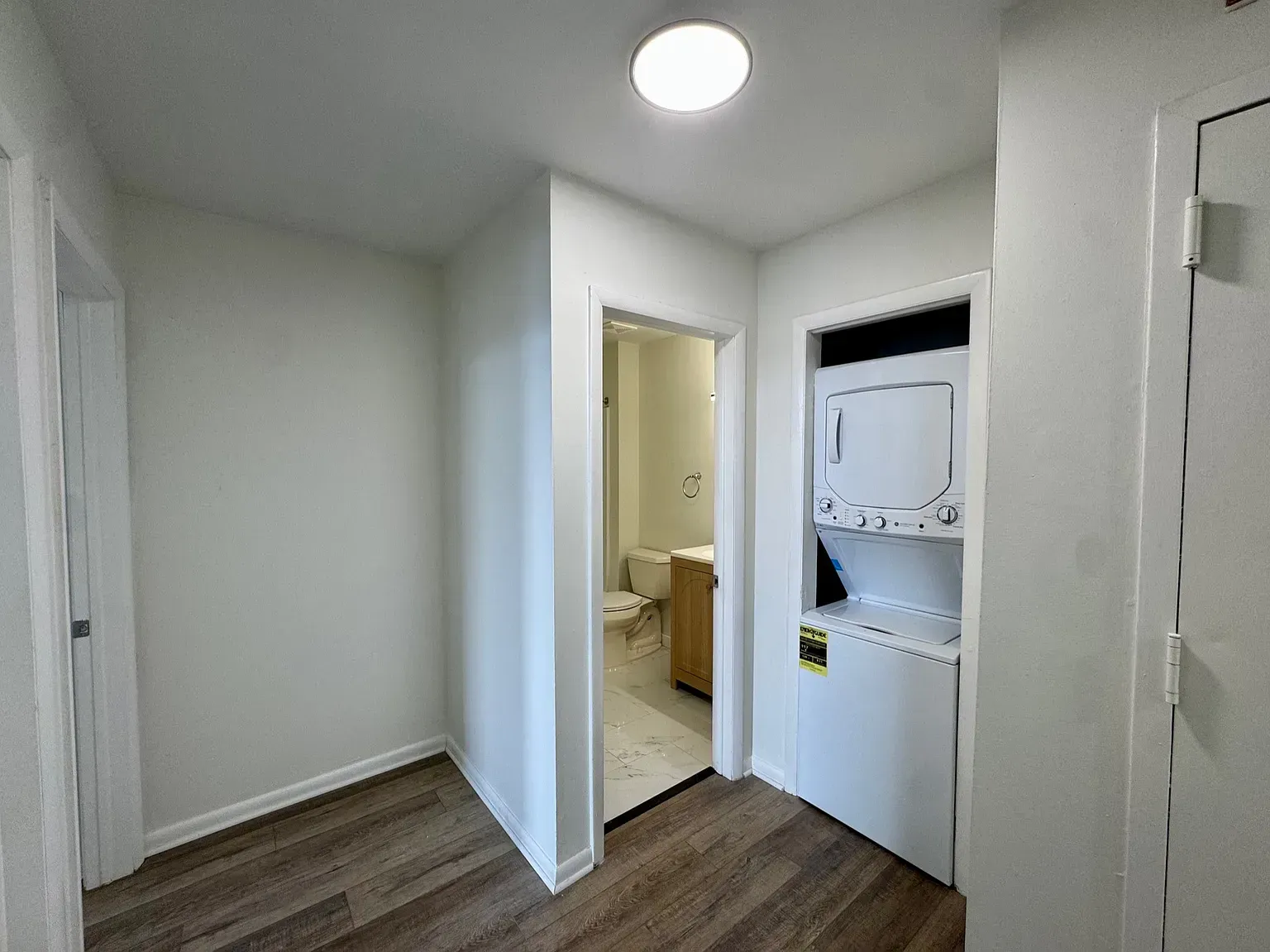 Empty white hallway with wood floors, a ceiling light, and a laundry nook beside an open bathroom door