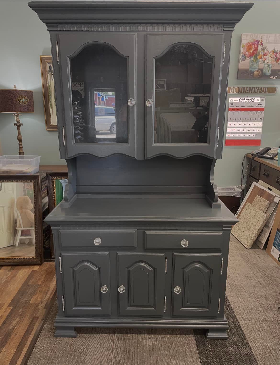 Gray painted hutch with glass doors, two drawers, and cabinet doors. Set in a room with a wooden floor.