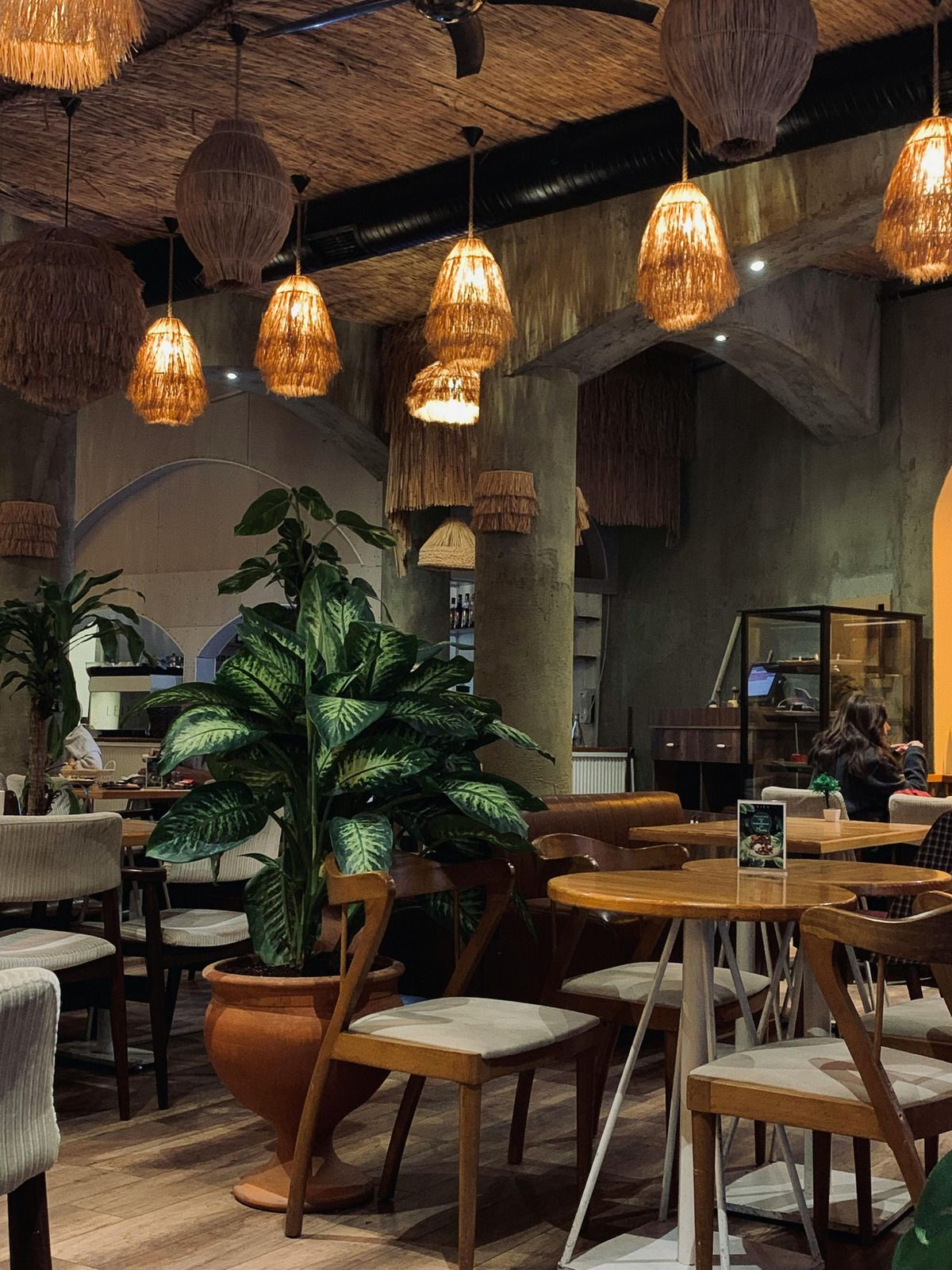 Cozy cafe interior with hanging woven lights, wooden tables and chairs, and large potted plant.