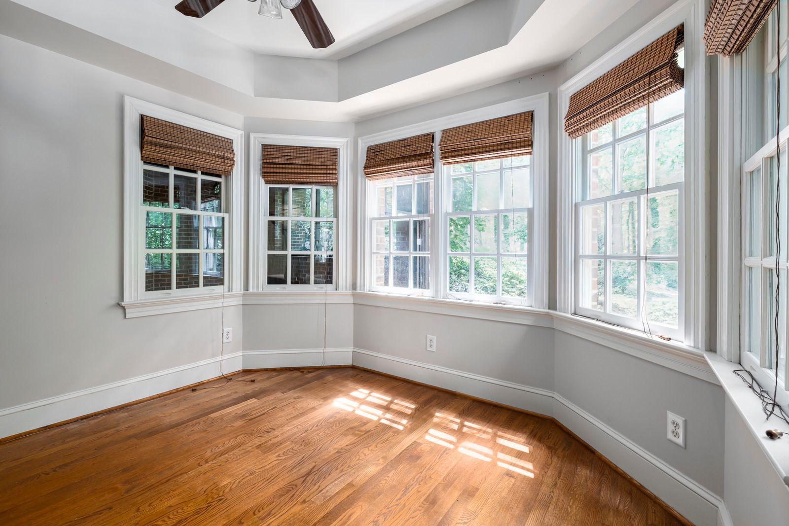 Empty sunlit room with hardwood floors, multiple windows, light gray walls, and brown roman shades.