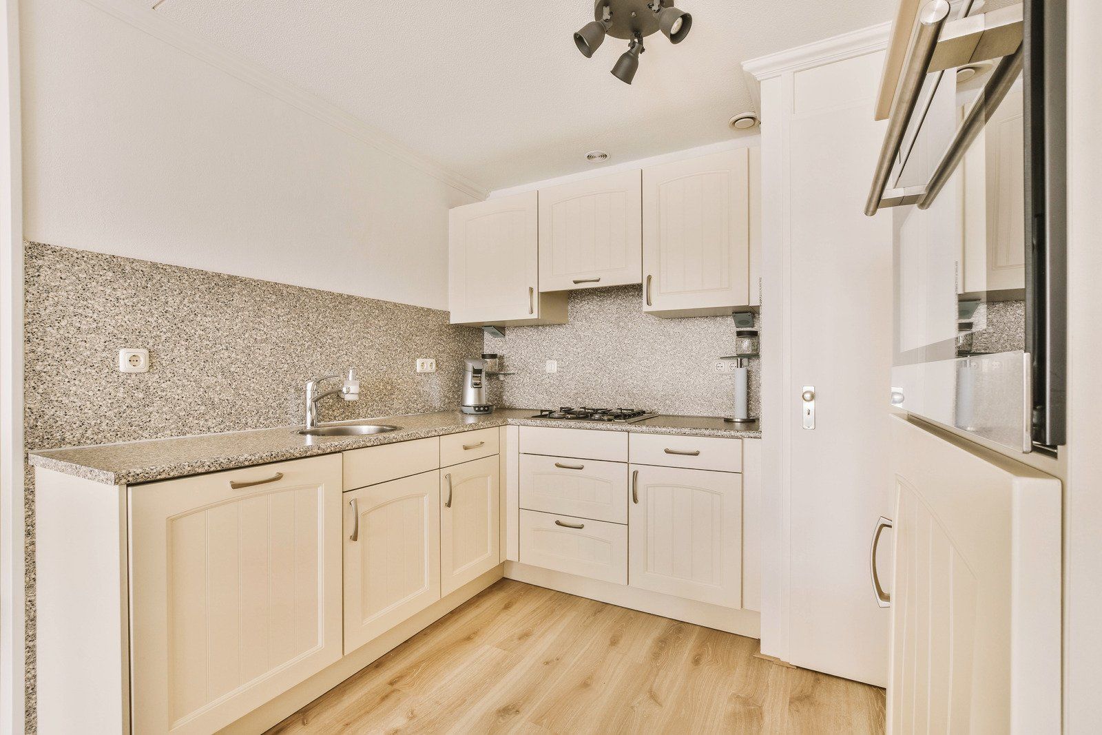 Off-white kitchen with light wood floors, granite countertops, and white cabinets; built-in oven and recessed lighting.