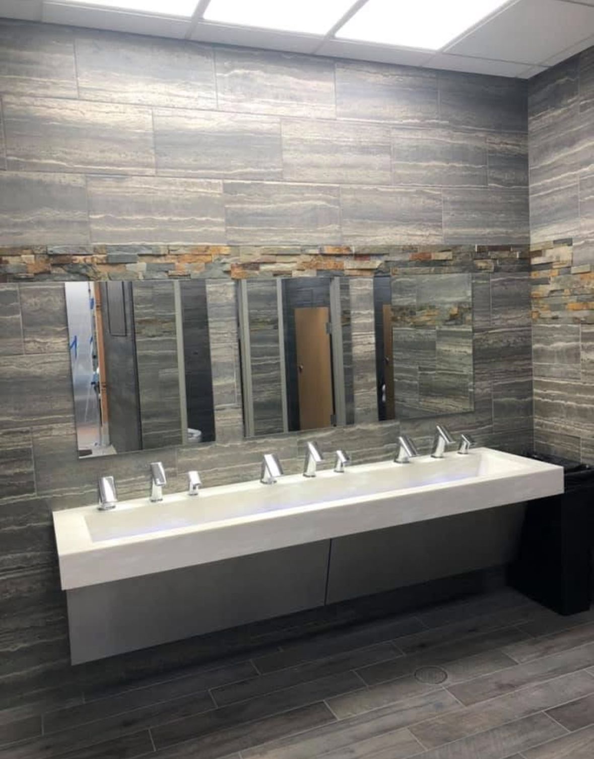 Public restroom with long white sinks, mirrors, gray tiled walls, and a black trash can.