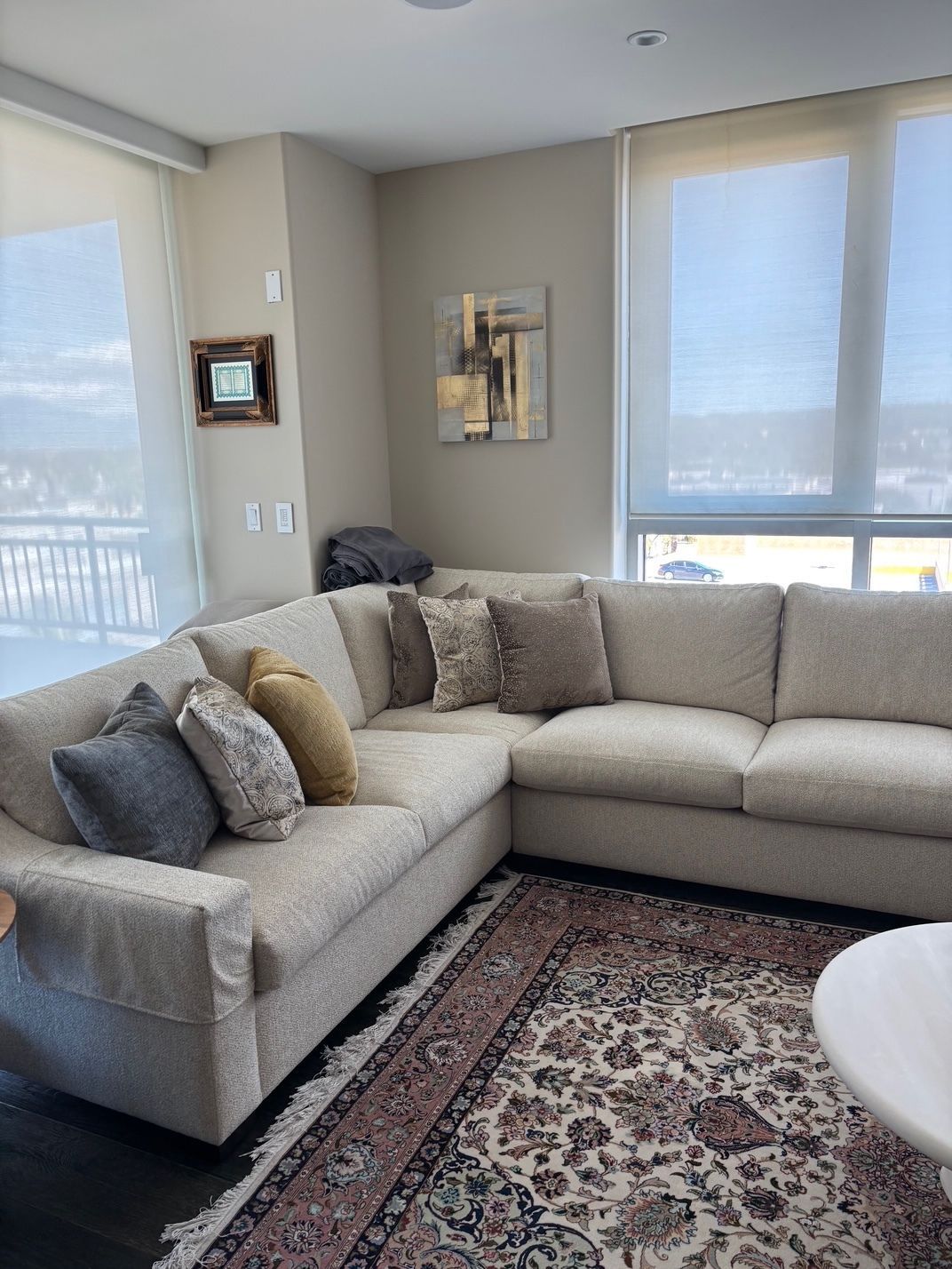 Beige sectional sofa with decorative pillows on an oriental rug near two large windows.