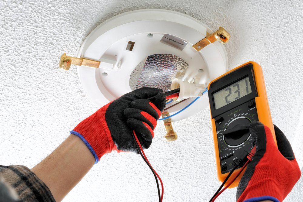 Electrician testing ceiling light fixture with a multimeter, wearing gloves.
