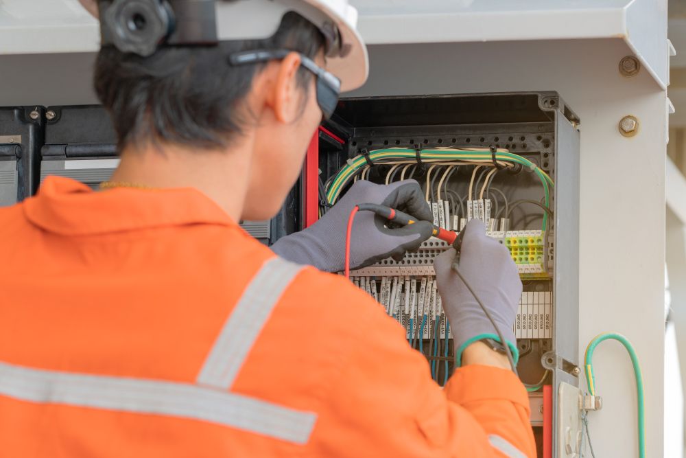 An electrician in orange coveralls tests wiring inside an electrical panel.