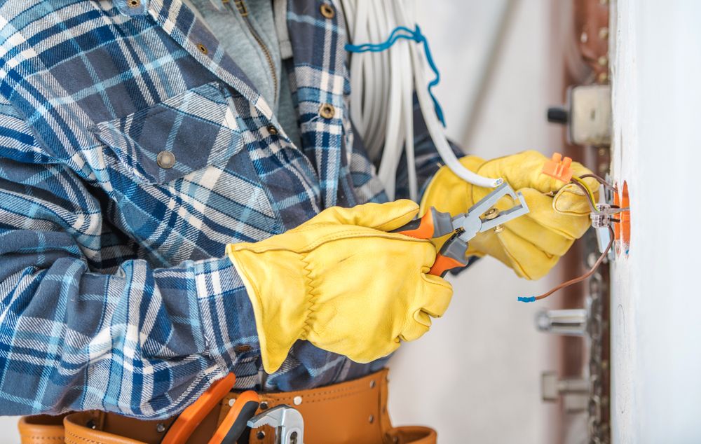 Electrician in yellow gloves wiring an outlet, wearing a plaid shirt.