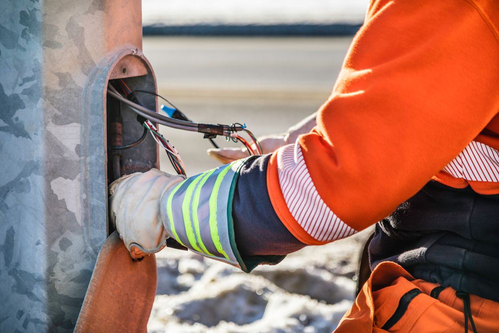 Person in orange workwear connecting wires inside a metal pole. Gloves, reflective stripes visible.