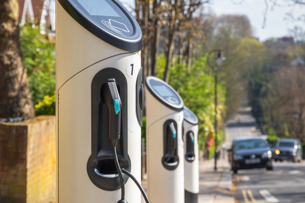 Electric vehicle charging stations on a street; cream-colored, with black and teal cables. Cars in the background.