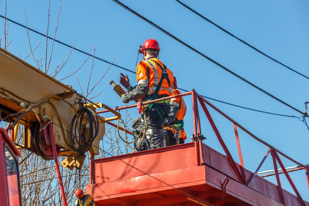 Lineman in a lift bucket working on power lines, wearing a red helmet and orange vest, blue sky.