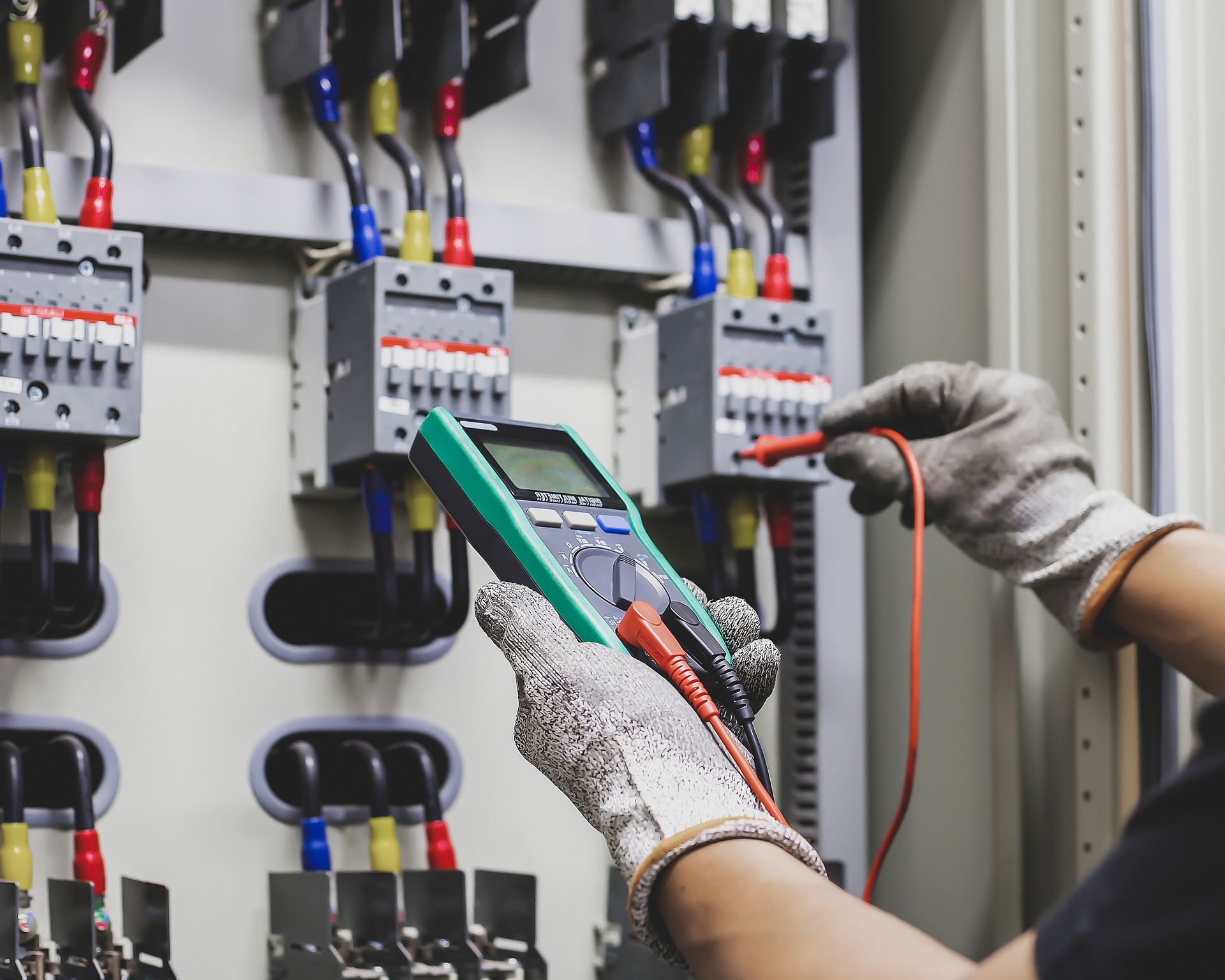 Electrician using a multimeter to test electrical components in a panel, wearing gloves.