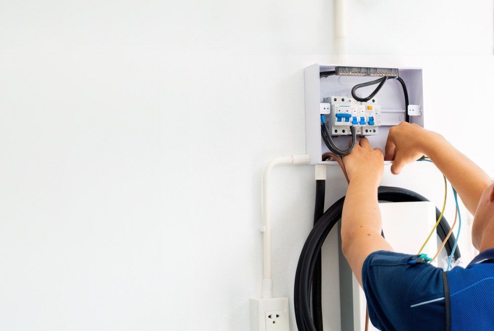 Electrician working on a circuit breaker box mounted on a white wall; blue shirt, black cable.