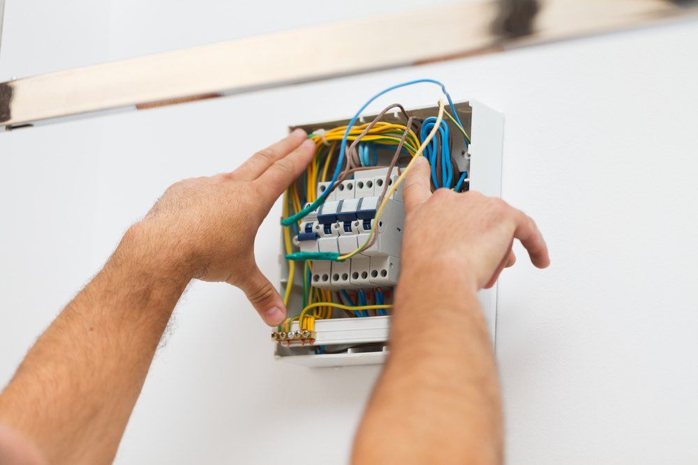 Person working on an electrical circuit breaker box, hands reaching, white wall.
