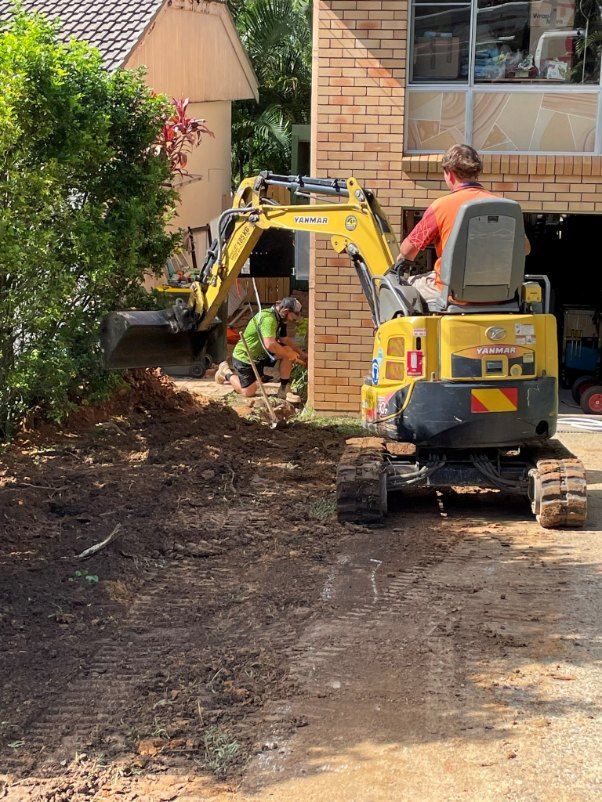 Workers using an excavator to dig in front of a house. One worker operates it, another near the dig site.