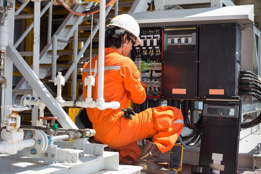 An electrician in orange overalls and a hard hat works on electrical equipment outdoors.