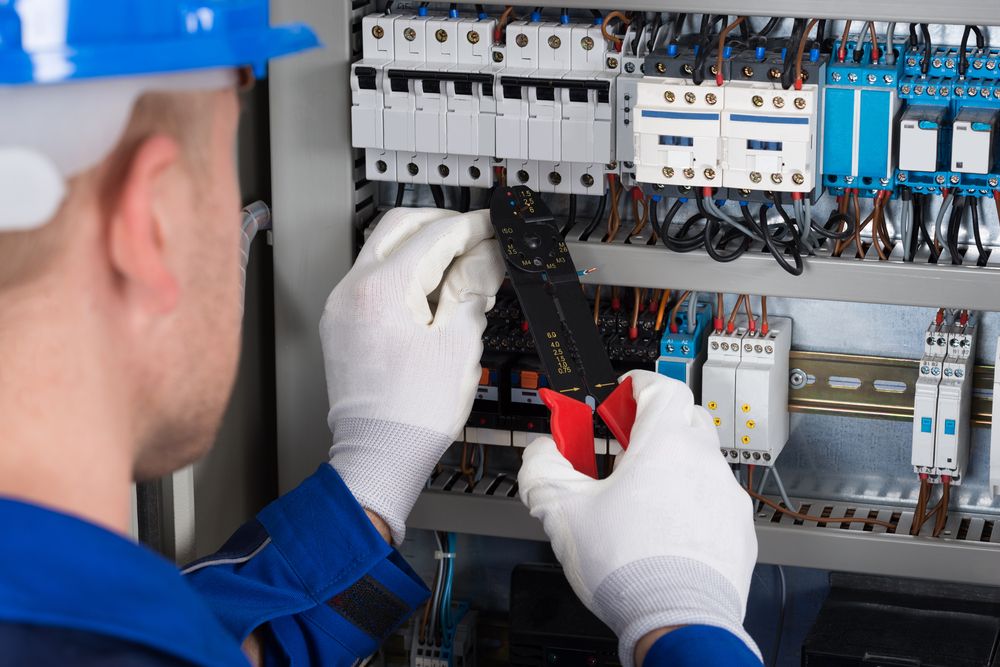 Electrician in blue coveralls and hard hat working on electrical panel, using wire strippers.