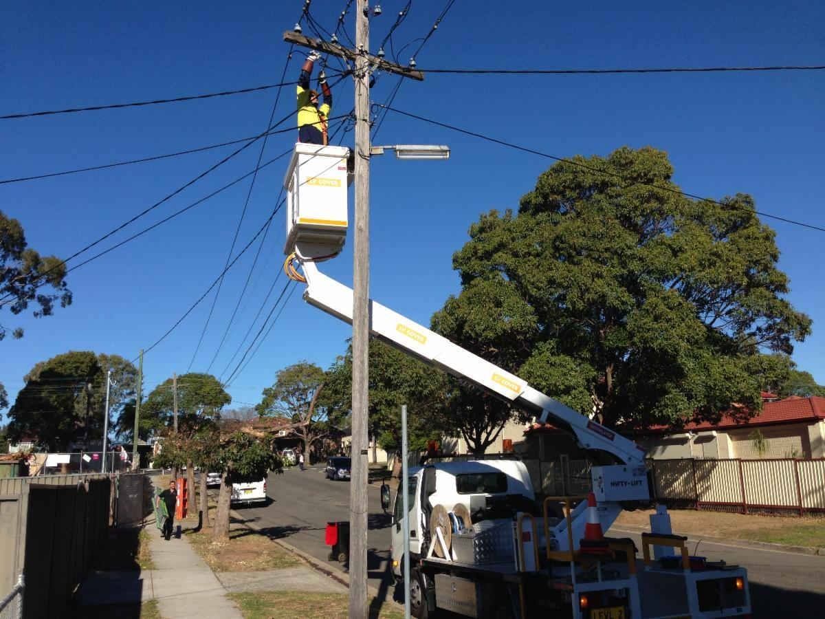 Lineman in bucket truck working on power lines on a sunny street with houses and trees.