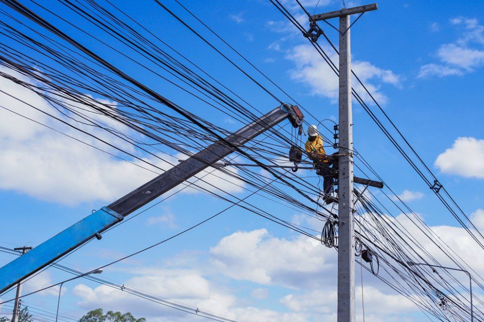 Lineman on a utility pole in a lift, working on a tangle of power lines against a blue sky with clouds.