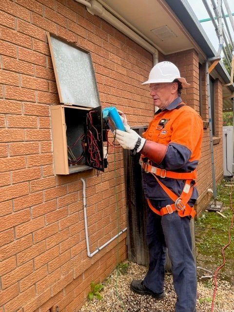 Electrician in safety gear working on a wall-mounted electrical box, using a blue tool. Brick building exterior.