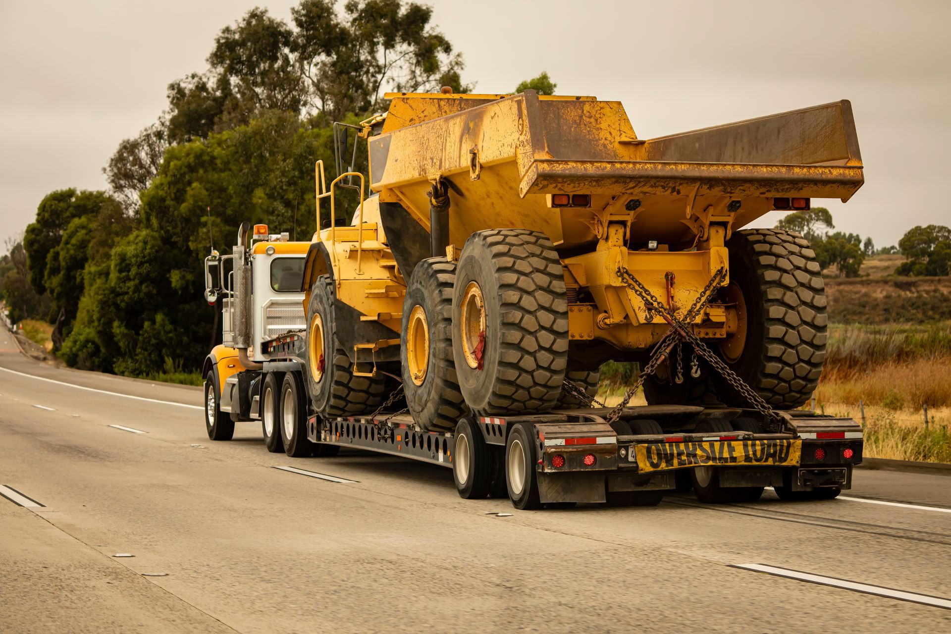 Oversize load semi truck hauling large yellow mining dump truck on flatbed trailer on highway.