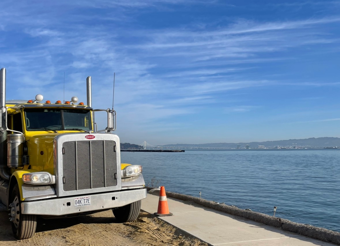 Yellow semi-truck parked near a shoreline. Water and distant hills under a blue sky.