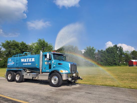Blue water truck spraying water, creating a rainbow, against a blue sky.
