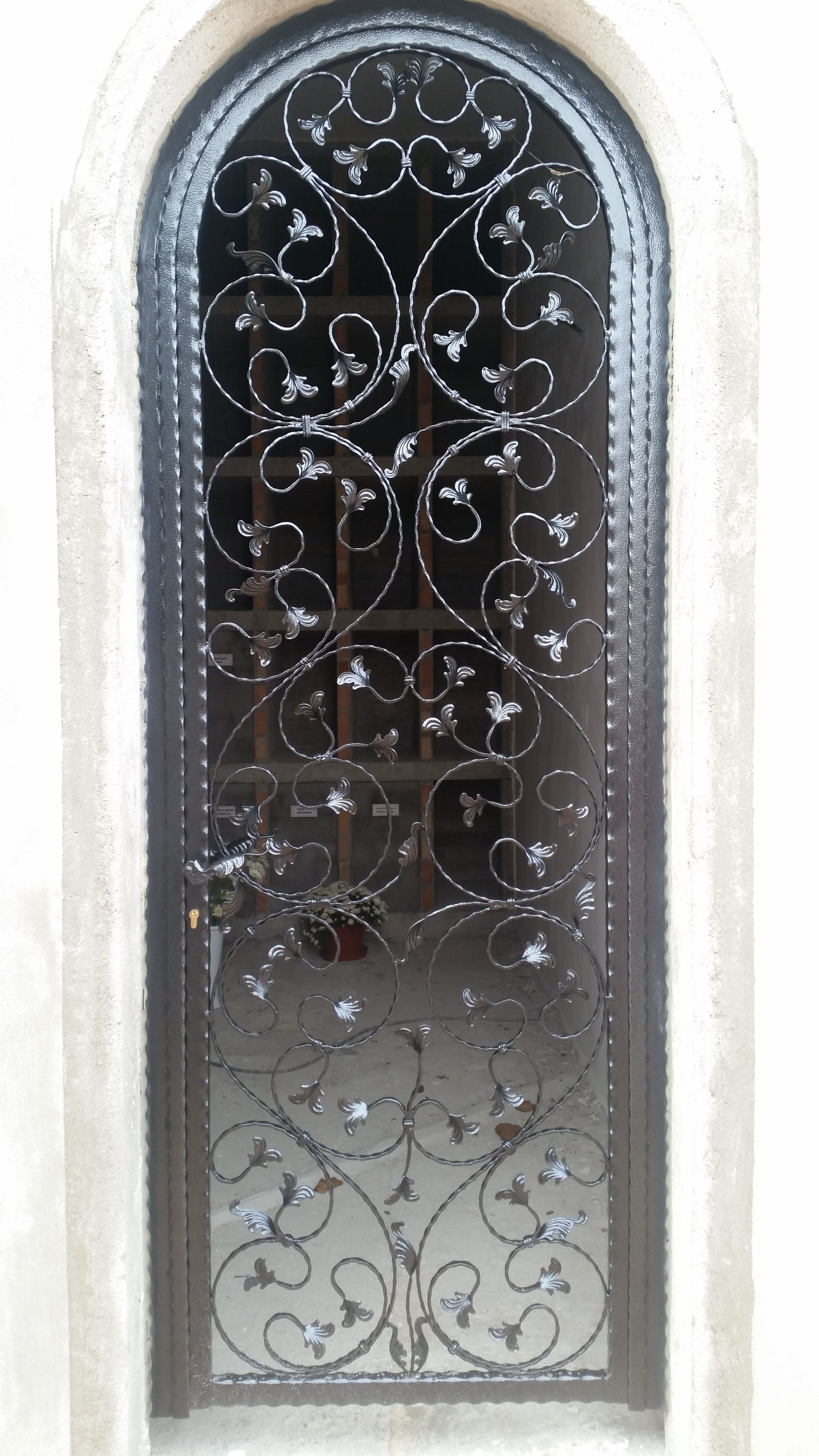 Cancello ad arco decorativo in ferro battuto con elaborate decorazioni a volute, inserito in un portale in pietra chiara.