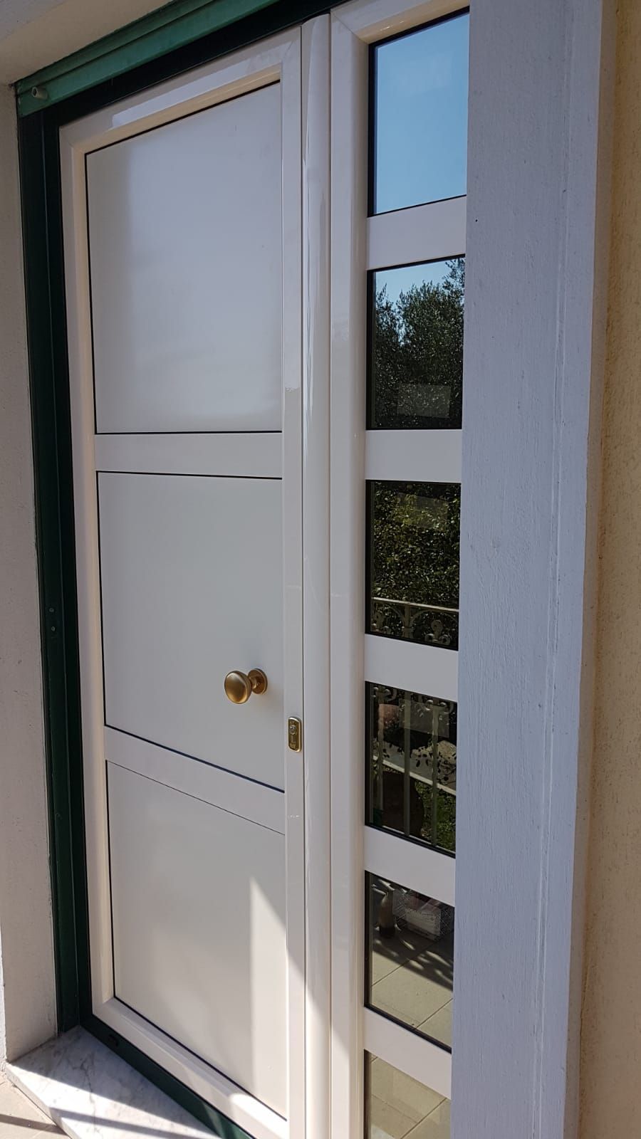 Porta d'ingresso bianca con pomello in ottone accanto a una stretta finestra verticale su una parete esterna soleggiata.