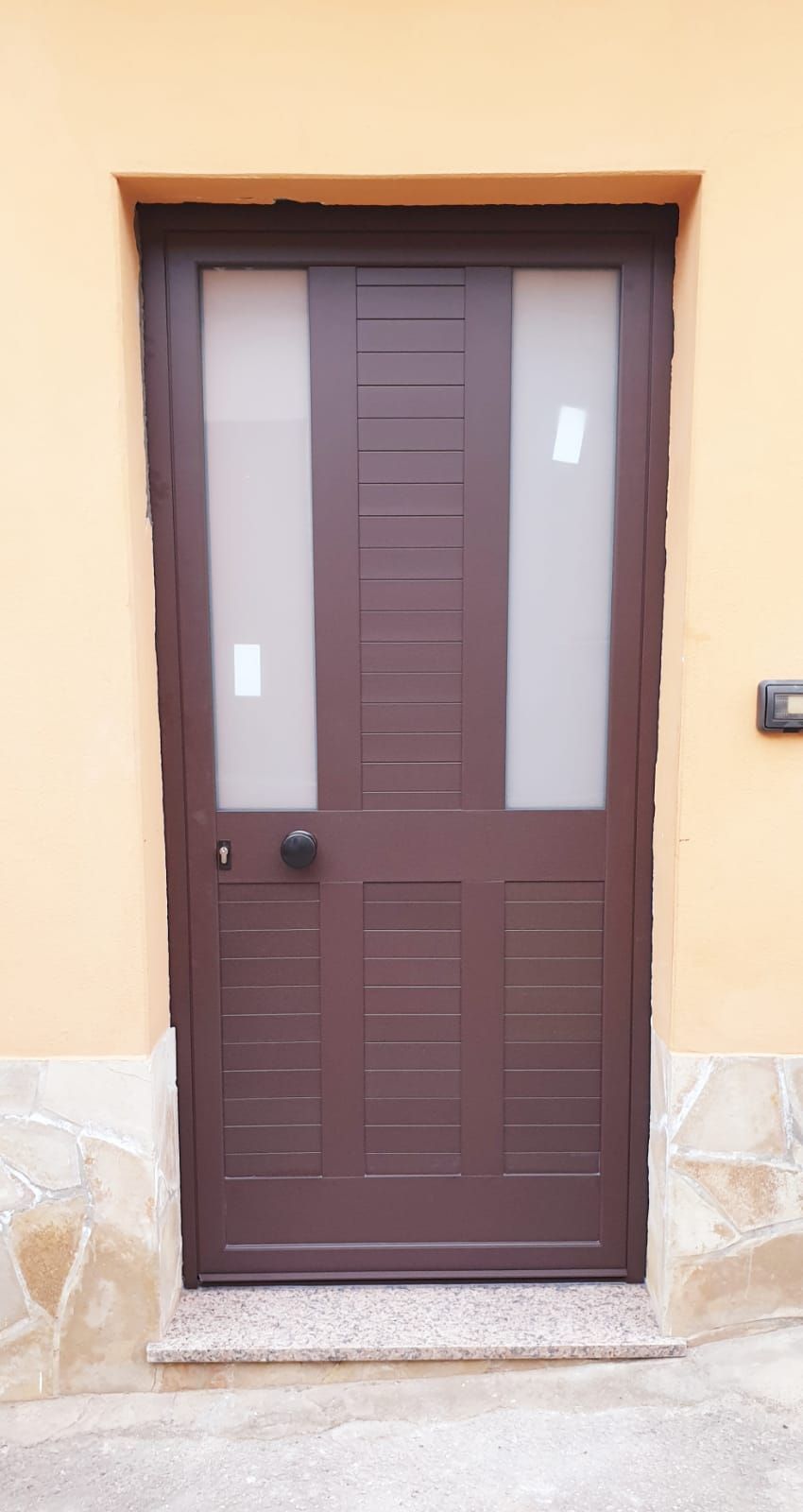 Porta d'ingresso marrone con pannelli in vetro satinato incastonati in una parete esterna beige.