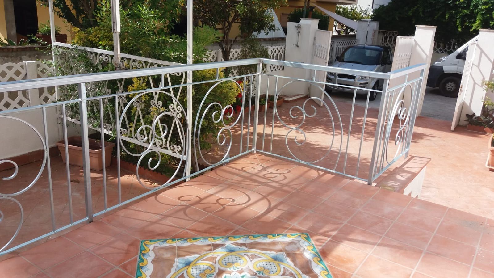 Terrazza con ringhiera in ferro battuto bianco, piastrelle in terracotta, piante in vaso e una sedia in pieno sole.