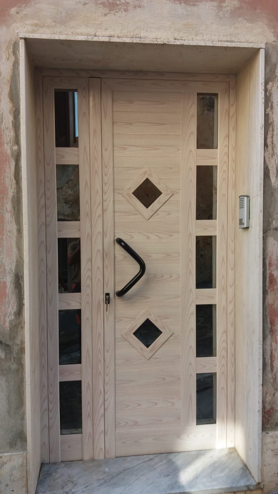 Porta d'ingresso in legno con finestre a rombo e finestre laterali verticali in una parete beige
