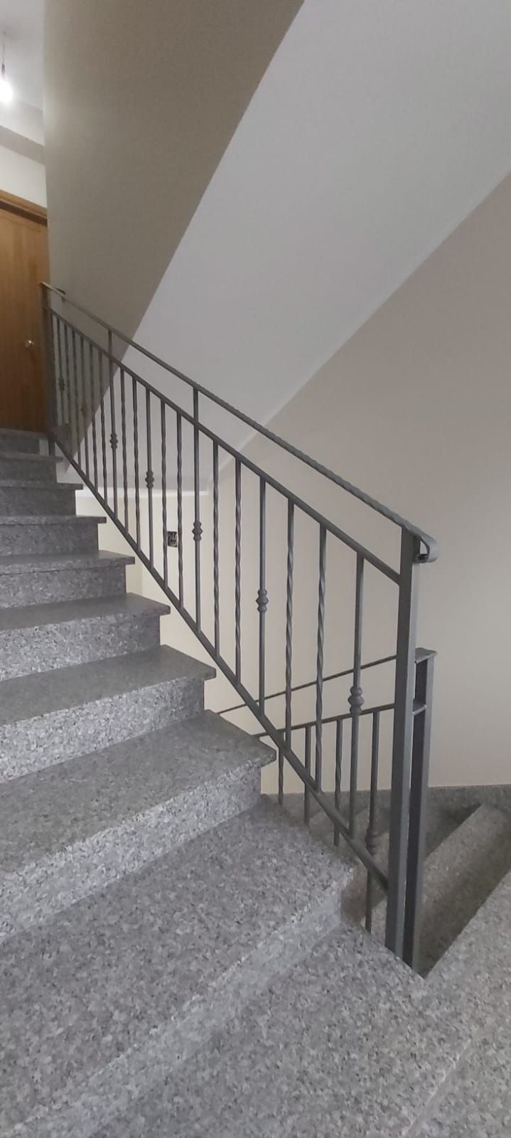 Vano scale interno con gradini rivestiti di moquette grigia e corrimano in metallo lungo una parete beige.