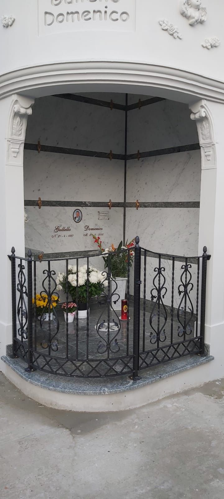 Piccola nicchia commemorativa bianca con ringhiera in ferro nero e fiori, con la scritta 